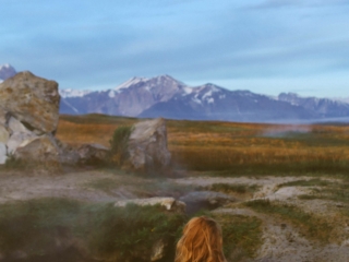 Woman in rainbow swimsuit sitting in a hot spring with mountains in the background